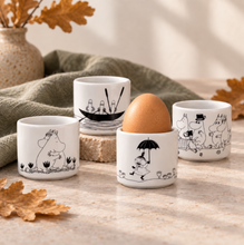 Load image into Gallery viewer, Four white ceramic egg cups with black Moomin-themed line drawings, arranged on a stone surface with a green cloth, leaves, and a vase in the background. One cup holds a brown egg.