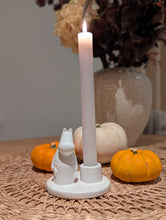 Load image into Gallery viewer, A white ceramic candle holder shaped like a seated cat, holding a lit white taper candle, styled on a woven placemat with small pumpkins and a vase in the background.