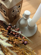 Load image into Gallery viewer, A close-up of a white ceramic candlestick holder with a white candle inserted, styled on a wooden surface alongside dried grasses, small purple berries, and a decorative house ornament.