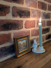 Load image into Gallery viewer, A lit teal pillar candle in a white ceramic Moomin character holder, styled on a wooden mantel next to a small framed landscape print against a red brick wall.