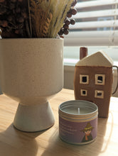 Load image into Gallery viewer, A small round tin candle with a lavender label and cartoon illustration sits on a wooden table in warm sunlight, next to a speckled cream-colored ceramic planter with dried flowers and a brown ceramic house-shaped object.