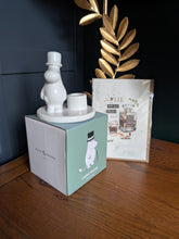 Load image into Gallery viewer, A white ceramic Moomin-themed candle holder set with a character figure and small cup on a round base, displayed next to its mint green and white 'Clammy Design' box and a printed card on a wooden tabletop.

