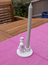 Load image into Gallery viewer, A white candle holder featuring a small Moomin character figurine and a tall taper candle, displayed on a pink tablecloth outdoors on a wooden table.

