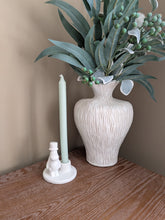 Load image into Gallery viewer, A white ceramic candle holder with a small snowman figure and a tall pale green candle, placed beside a large ribbed white vase filled with eucalyptus branches and green berries on a wooden surface against a beige wall.
